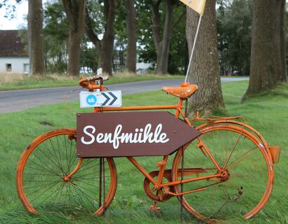 Ein orangenes Fahrrad mit einem Holzschild auf dem "Senfmühle" steht und nach rechts zeigt.