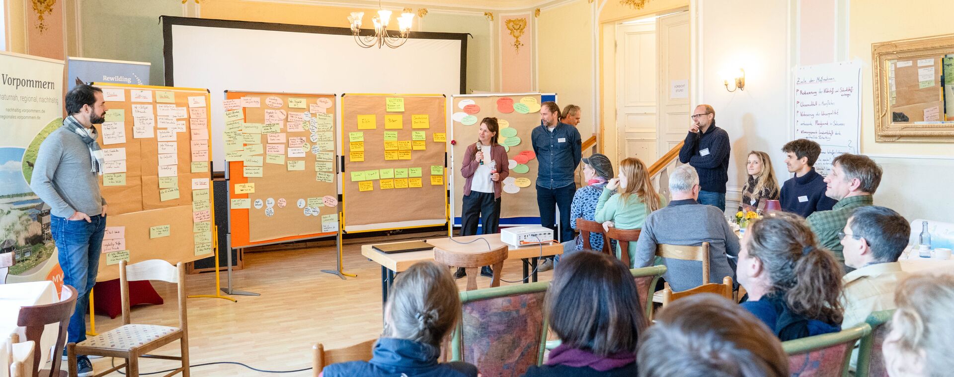 Stakeholder Workshop - Rewilding Oder Delta e.V Eine Gruppe an Menschen hört aufmerksam zwei Moderatoren zu, die die Ergebnisse eines Workshops präsentieren