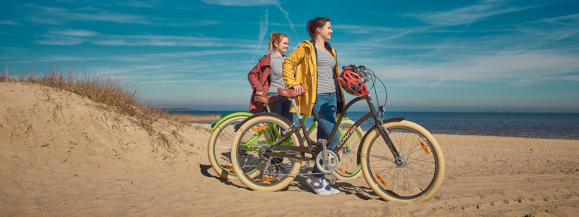 There are two women on the beach who push their bikes through the sand. On the left is a small dune and the water in the background.