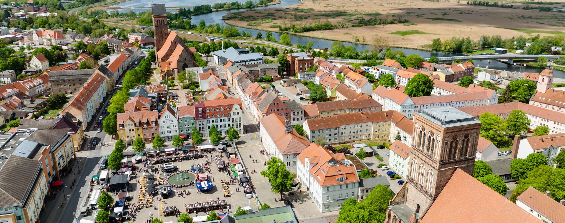 Eine Luftaufnahme über den Marktplatz in Anklam, auf dem reges Treiben herrscht und dahinter die Peene