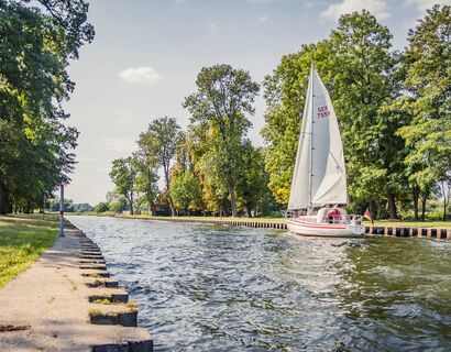 Einfahrt Richtung Hafen eines Segelbootes. Links und rechts sind Bäume und die Uferkante befestigt.