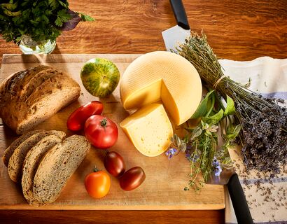 Eine Platte mit einem runden Laib Käse, daneben Tomaten und ein Brot. Rechts vom Käse liegen Kräuter und Lavendel