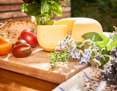 Eine Platte mit einem runden Laib Käse, daneben Tomaten und dahinter Brot. Im Vordergrund liege noch einige Kräuter