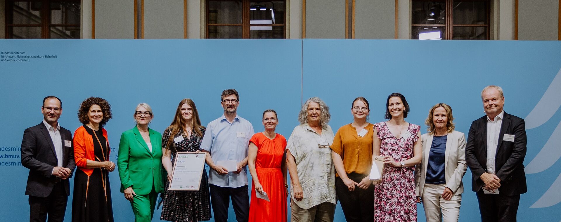 Several people stand side by side and smile in the camera, two hold the certificate and price in the camera