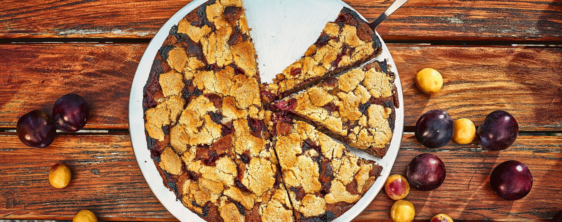 A little more than three quarters of a plum cake from above. There is a cake lifter under one piece and plums are lying around the cake.