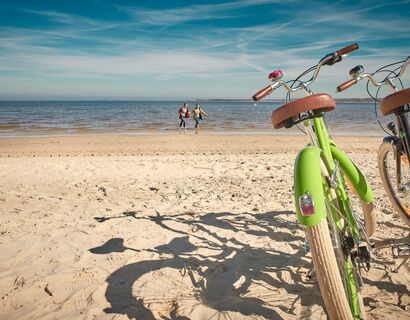 On the right in the picture there is a green and brown bike in the sand from the beach. Two people run through the water in the background.