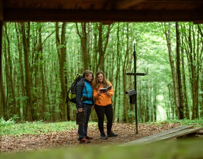 Zwei Wanderinnen, aus einem Rahmen heraus fotografiert, stehen an einem Kreuz und lesen etwas aus einem Buch