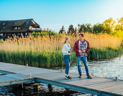 A man and a woman stand on a jetty, the Haffhus Ueckermünde can be seen in the background