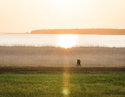 Luftaufnahme eines Spaziergangs von zwei Personen. Im Hintergrund Schilf und Wasser, während die Sonne aufgeht.