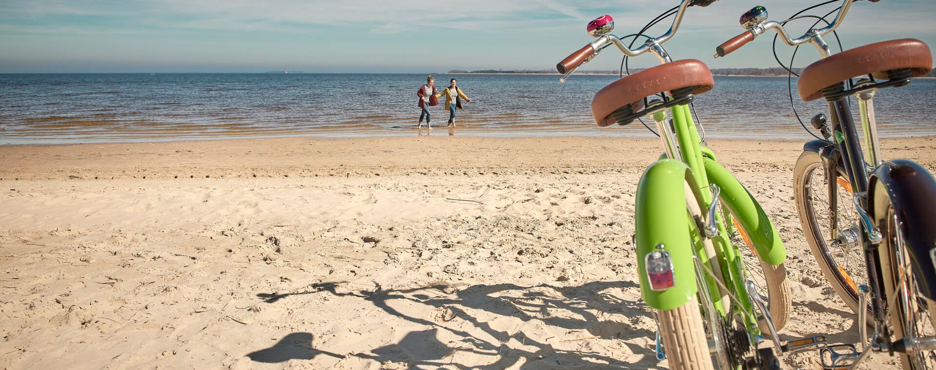 On the right in the picture there is a green and brown bike in the sand from the beach. Two people run through the water in the background.