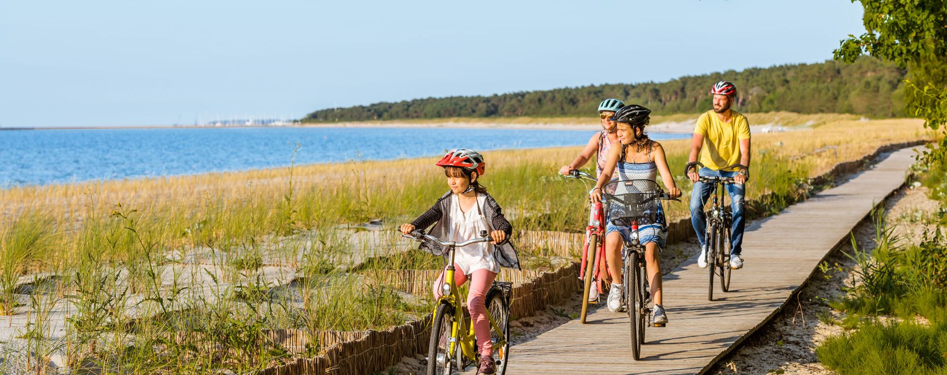 Family runs with bicycles over wood on the Lubminer beach. On the left is the beach and the water, there are trees on the right. The family looks towards the beach.