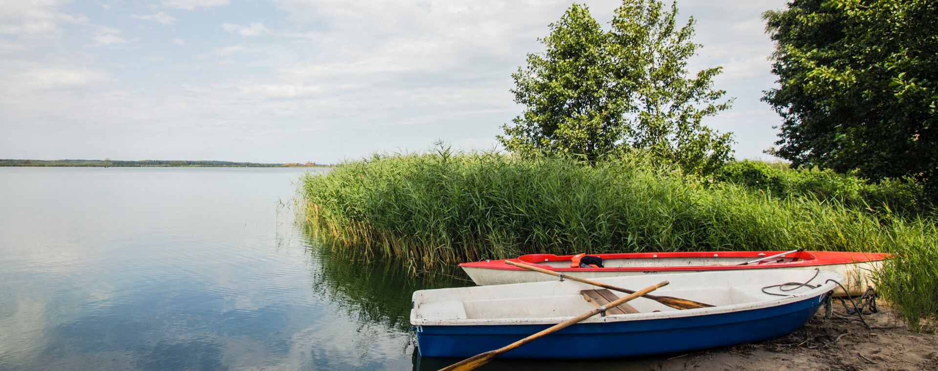 Zwei Ruderboote liegen halb an Land und halb im Wasser. Eins ist blau, das hintere ist weiß-rot. Im Hintergrund Schilf und Wasser.