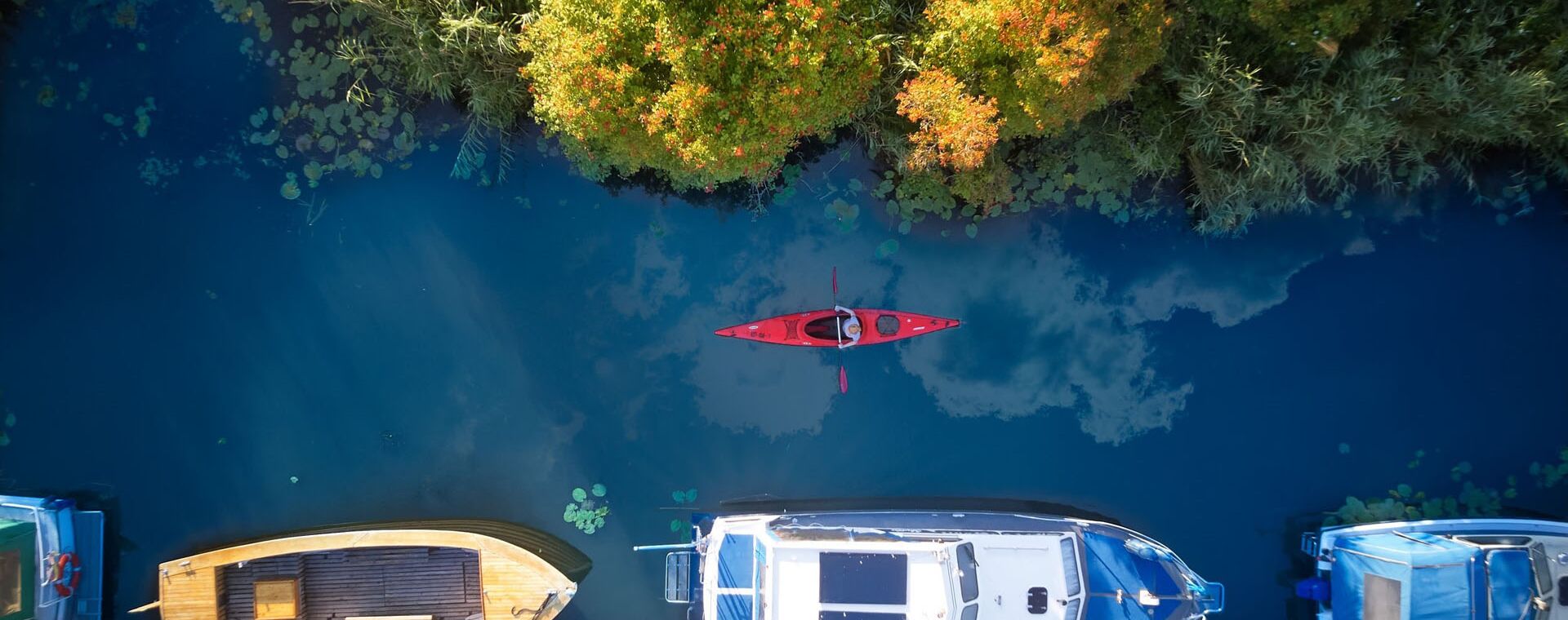 Aufnahme aus der Vogelperspektive, von links nach rechts sieht man eine Fluss mit verschiedenen Booten am unteren Hafenanleger , auf dem Fluss fährt ein Kanu und oben ist Wald