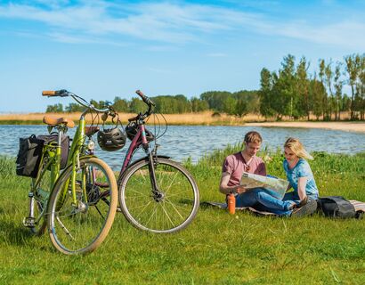 Links im Bild zwei Fahrräder, rechts daneben sitzt ein Paar auf einer Decke im Gras und guckt auf eine Karte im Hintergrund Wasser und Strand.