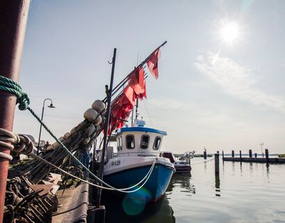 Ein Blick aus dem Hafen Altwarp. Vorne im Bild sind rote Fahnen angelehnt und ein Fischerboot liegt im Hafen