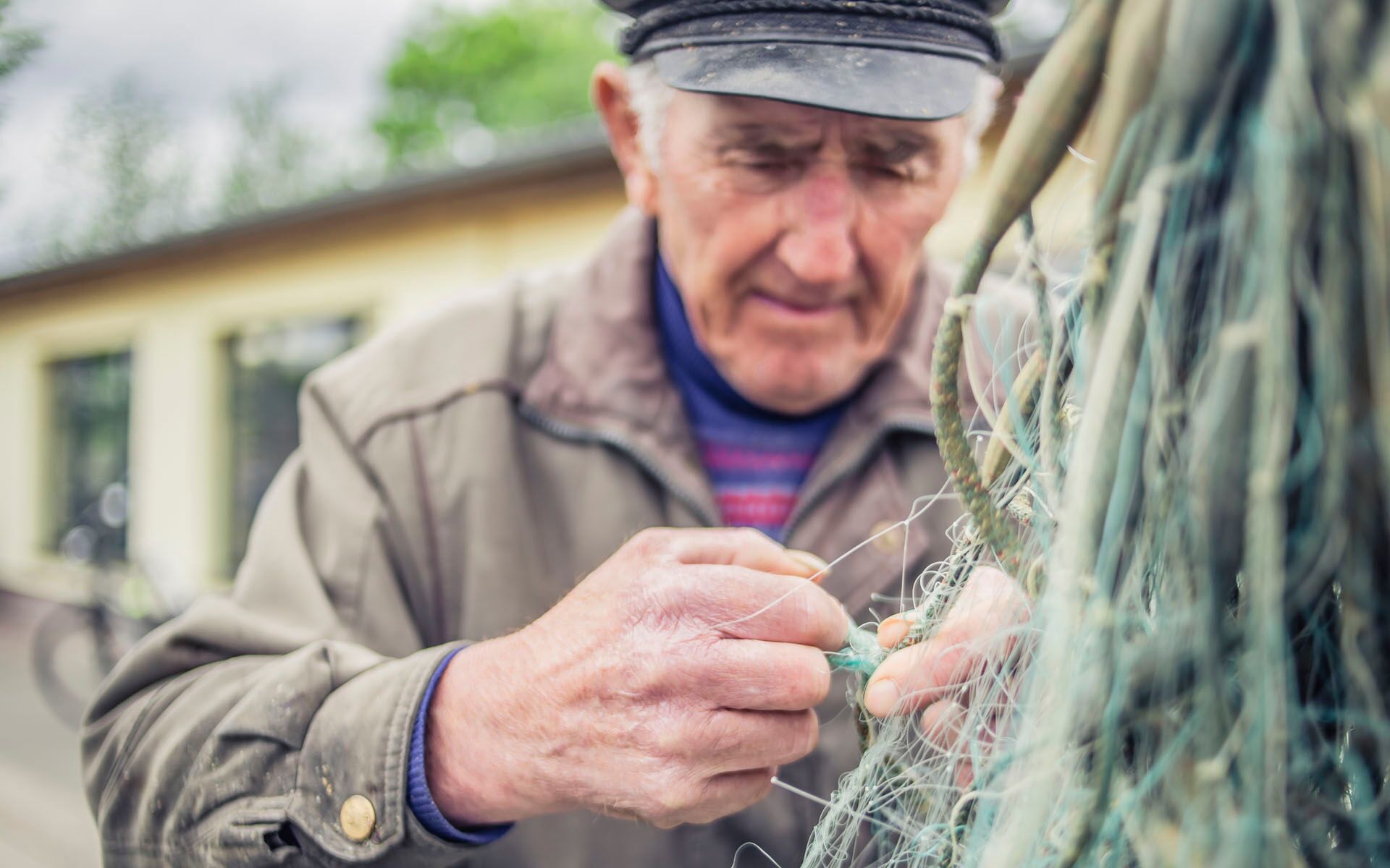 Ein älterer Herr mit einer Art Schiebermütze von vorne fotografiert, wie er ein Fischernetz ausknotet.