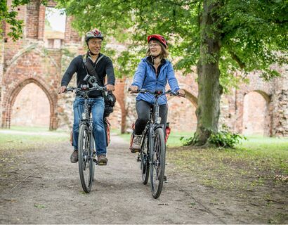 Zwei Fahrradfahrer fahren den Weg von der Klosterruine weg. Im Hintergrund Ausschnitt der Klosterruine Eldena sichtbar.