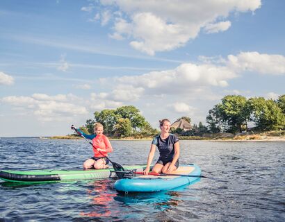 Two women kneel on SUPs on the water, with the beach in the background.