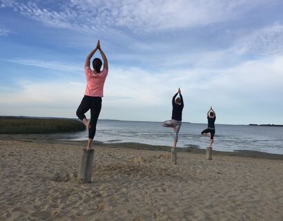 Drei Personen stehen auf Holzstumpen in der Baum-Position am Strand. Sie gucken alle Richtung Wasser.