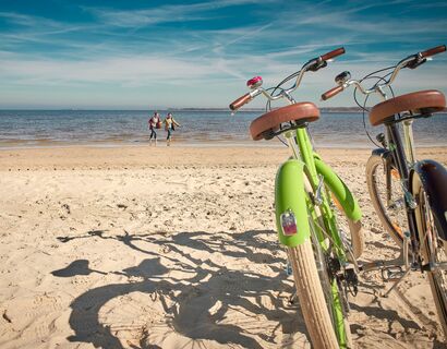 Freest Strand Rechts im Bild stehen ein grünes und braunes Fahrrad im Sand vom Strand. Im Hintergrund laufen zwei Personen durchs Wasser.