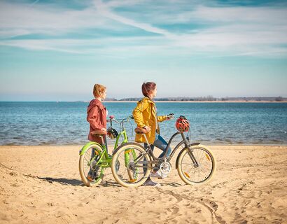 There are two women on the beach who push their bikes through the sand. The water in the background.