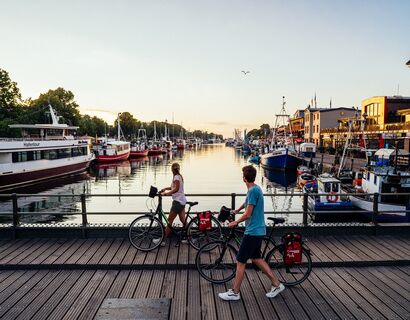 Radwandern entlang der Ostseeküste Zwei Radfahrer schieben ihr Rad über eine Brücke in Warnemünde in der Abenddämmerung und blicken aufs Wasser. Auf dem Wasser sind viele Boote.