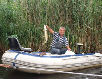 Angler, with an elongated fish in hand. He sits in a white rubber boat that is in front of reeds