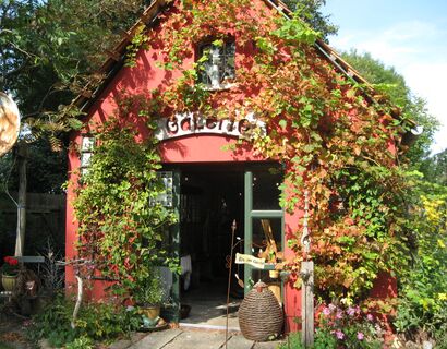 kleines rotes Haus mit der Überschrift Galerie, das Haus ist stark bewachsen und detailvoll dekoriert, die Tür steht offen bei gutem Wetter